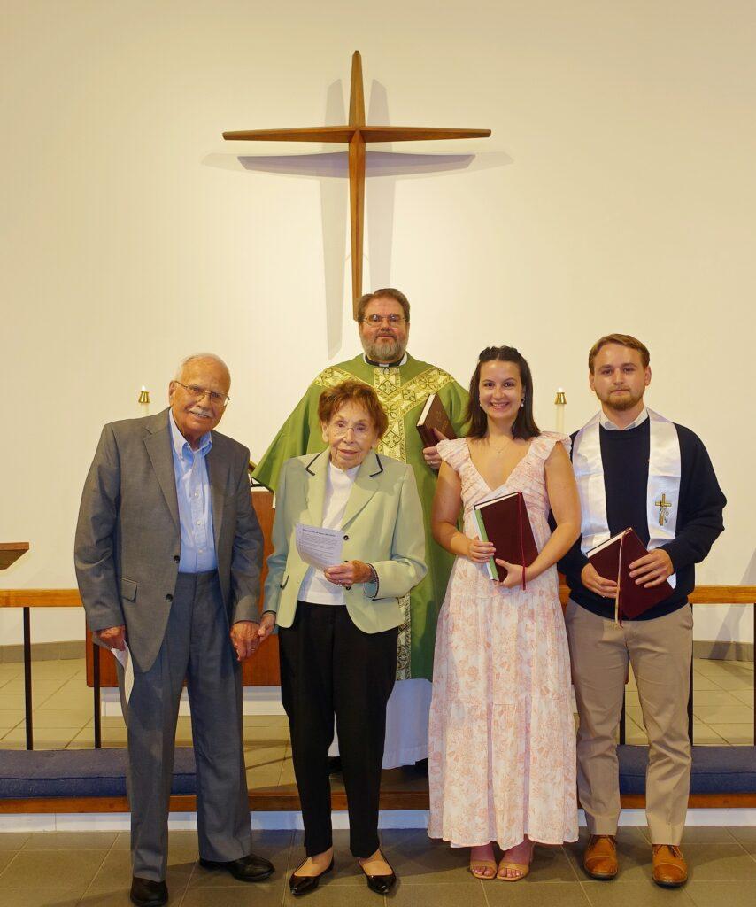 Five people stand inside a church before a large wooden cross mounted on the wall. A clergyman in a green and gold vestment stands at the center back, while four individuals stand in front of him. The two people on the left, an elderly man and woman, are dressed in formal attire; the woman holds a printed program. The two on the right, a younger woman in a light pink dress and a young man wearing a dark sweater with a white stole, each hold a Bible. The group appears to be participating in a church ceremony or recognition event.