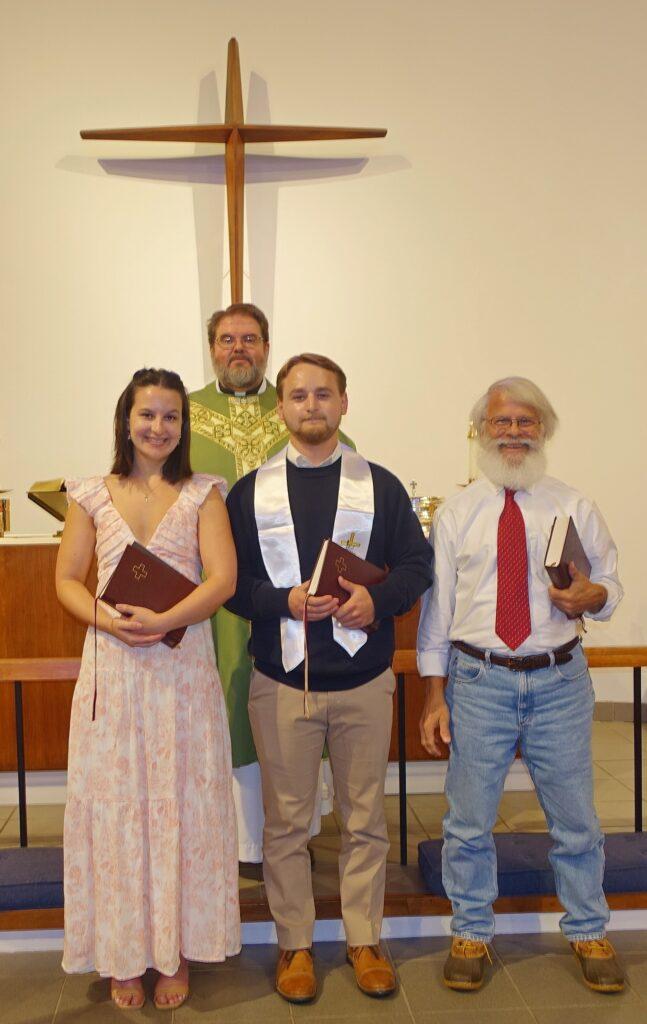 Four people stand inside a church before a large wooden cross mounted on the wall. A clergyman in a green and gold vestment stands behind three individuals holding Bibles. The woman on the left wears a light pink dress, the man in the center wears a dark sweater with a white stole, and the man on the right wears a white shirt with a red tie and jeans. The group appears to be participating in a religious ceremony or recognition event.