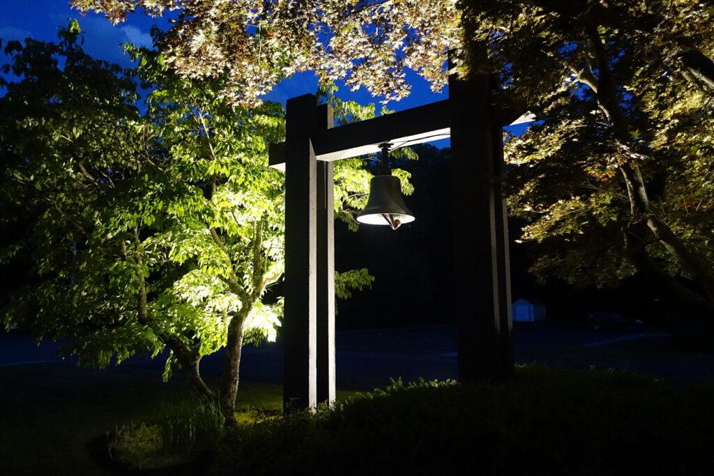 Illuminated wooden bell tower at night with a bronze bell hanging in the center, surrounded by spotlit green and autumn-colored trees under a deep blue evening sky.