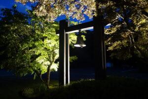 Illuminated wooden bell tower at night with a bronze bell hanging in the center, surrounded by spotlit green and autumn-colored trees under a deep blue evening sky.