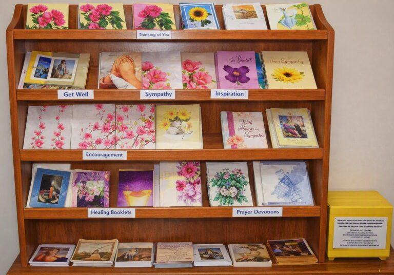 A wooden display rack with four shelves filled with colorful greeting cards and small booklets. Each shelf has labeled sections such as “Get Well,” “Sympathy,” “Inspiration,” “Encouragement,” “Healing Booklets,” and “Prayer Devotions.” The cards feature floral and inspirational designs. A yellow donation box sits to the right, and the photo has a warm tone.