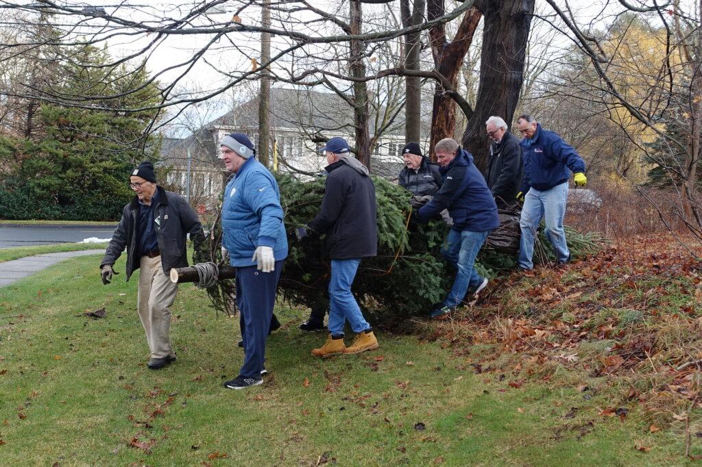 Seven men work together outdoors to carry a large evergreen tree trunk across a grassy area. They are dressed in winter clothing—jackets, hats, gloves, and boots—suggesting cold weather. The background shows bare trees, a residential street, and houses, indicating late autumn or early winter.