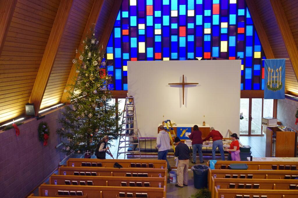 Inside a church sanctuary with a high wooden ceiling and vibrant blue, red, and yellow stained-glass windows, several people decorate a tall Christmas tree beside the altar. The altar features a wooden cross on a white wall and a blue cloth with a gold angel design. Volunteers work together arranging decorations and supplies, creating a warm, festive atmosphere.