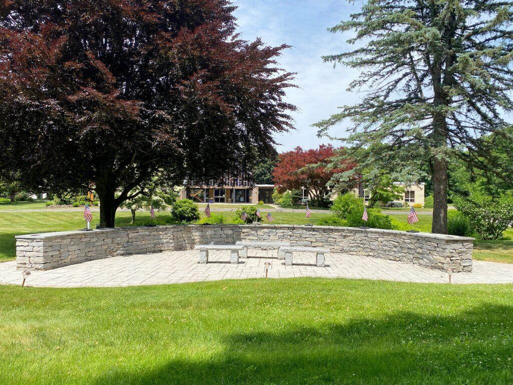 A peaceful outdoor view of the Cheshire Lutheran Church Columbarium surrounded by lush green lawns and mature trees. The curved stone wall encloses a paved area with benches, and small American flags line the top of the wall. In the background, the church building is partially visible among trees with deep red and green foliage under a bright blue sky.