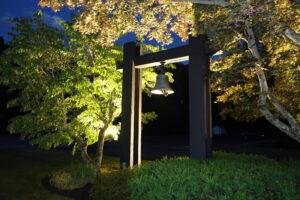 Outdoor bell mounted on a dark wooden frame, illuminated by warm lights at night. The structure is surrounded by lush green and reddish trees, their leaves glowing under the lighting against a deep blue evening sky. The scene feels peaceful and well‑maintained, highlighting the bell as the focal point among the foliage.