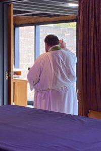 Alt text: A pastor dressed in a white robe with a green stole stands near an open doorway, raising his hand in a gesture of blessing or prayer. The scene is softly lit, with natural light coming through large windows that reveal a brick wall outside. In the foreground, a table covered with a deep purple cloth is visible, suggesting a church or ceremonial setting.