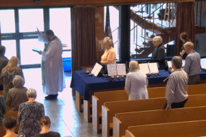 A church service scene showing a pastor in a white robe with a green stole standing at the front, raising his hand in blessing while holding an open book. Congregants stand in pews facing forward, heads bowed. In front of them, several people stand behind tables covered with dark blue cloths displaying handbells and open sheet music, suggesting a handbell choir setup. Sunlight streams through large glass doors in the background, and an American flag and staircase are visible beyond the windows.