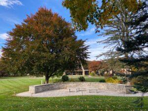 A peaceful outdoor view of the Cheshire Lutheran Church grounds featuring the stone Columbarium in the foreground. The circular stone wall encloses a paved area surrounded by lush green grass and autumn trees with red, orange, and green leaves. The church building with its distinctive A‑frame roof is visible in the background under a bright blue sky with scattered clouds.