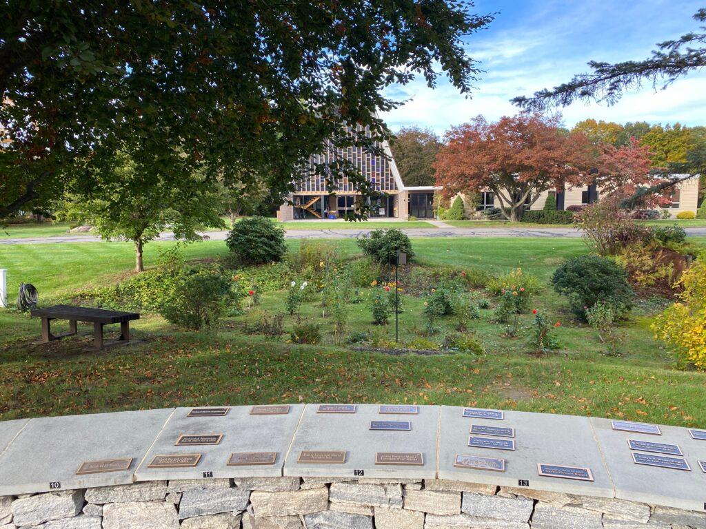 A peaceful view of the Cheshire Lutheran Church grounds from the Columbarium. Bronze memorial plaques line the curved stone wall in the foreground. Beyond them lies a small garden with blooming flowers, shrubs, and a wooden bench under a large tree. The church building, with its distinctive A‑frame roof and glass façade, stands in the background surrounded by autumn-colored trees and green lawns under a bright blue sky.