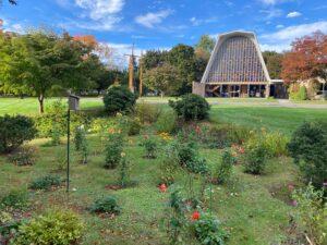 A vibrant autumn scene featuring the Cheshire Lutheran Church with its tall A‑frame roof and large glass façade. In the foreground, a memorial garden blooms with colorful flowers—red, yellow, orange, and pink—surrounded by green shrubs and trees. A small birdhouse stands on a pole near the garden, and three tall wooden crosses rise prominently on the lawn to the left. The sky above is bright blue with scattered white clouds.
