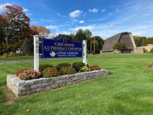 A bright autumn scene featuring the Cheshire Lutheran Church sign in the foreground, surrounded by colorful flowers and neatly trimmed greenery. The blue and gold sign reads “Cheshire Lutheran Church” and “Little Cherubs Christian Preschool,” with the number 660 displayed vertically on the left post. Behind the sign, the church building stands with its distinctive A‑frame roof and tall glass façade, flanked by trees with red, orange, and green leaves under a vivid blue sky.