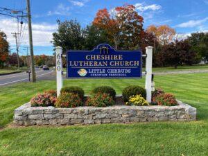 A blue and gold sign for Cheshire Lutheran Church stands on a stone base surrounded by colorful flowers and neatly trimmed greenery. The sign includes the words “Little Cherubs Christian Preschool” and the street number 660 displayed vertically on the left post. Behind the sign, a road curves through a tree-lined area with autumn foliage in shades of red, orange, and green under a bright blue sky.