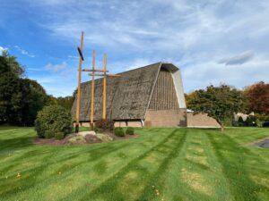 A wide view of the Cheshire Lutheran Church building with its steep, shingled roof and tall, narrow windows. Three large wooden crosses stand prominently on the manicured lawn in front of the church, surrounded by neatly trimmed shrubs and landscaped mulch beds. The sky above is partly cloudy with patches of blue, and autumn trees add warm tones of red and orange in the background.