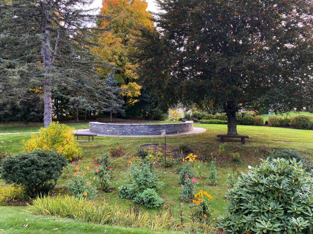 A tranquil garden scene showing the stone Columbarium surrounded by lush greenery and autumn trees. Benches sit beneath large trees offering shaded spots for reflection. In the foreground, colorful flowers bloom among shrubs and grass, while tall evergreens and golden foliage frame the background under a partly cloudy sky.