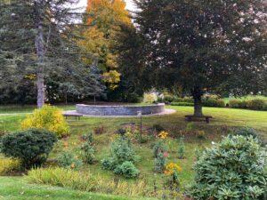 A tranquil garden scene showing the stone Columbarium surrounded by lush greenery and autumn trees. Benches sit beneath large trees offering shaded spots for reflection. In the foreground, colorful flowers bloom among shrubs and grass, while tall evergreens and golden foliage frame the background under a partly cloudy sky.