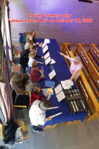A group of children stand along a long table covered with a dark blue cloth inside a church. Each child holds a handchime while looking at sheet music arranged in rows. An adult instructor stands on the opposite side of the table, pointing toward the music with a pencil. Wooden pews line the background, and sunlight filters through tall windows, creating a warm, inviting atmosphere.