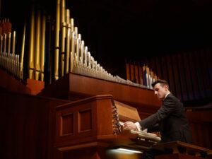 A young man in a dark suit plays a large pipe organ in a concert hall. The organ console is made of polished wood with multiple keyboards and rows of stop knobs. Behind him rises an impressive array of gold and silver organ pipes arranged in ascending height, illuminated warmly under stage lighting.