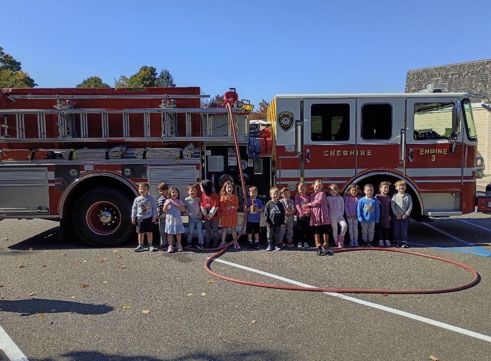 Children from Little Cherubs Christian Preschool stand in a line in front of a red Cheshire Fire Department truck, smiling during a visit. A fire hose lies coiled on the ground, and the bright blue sky and fall trees frame the cheerful scene.