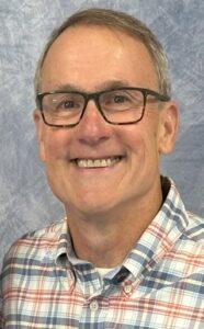 Portrait of the House and Grounds Deacon at Cheshire Lutheran Church, wearing glasses and a plaid button‑up shirt, smiling against a light blue textured backdrop.