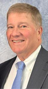 Portrait of the Financial Secretary at Cheshire Lutheran Church, wearing a dark suit, white shirt, and light blue tie, smiling against a soft blue studio backdrop.