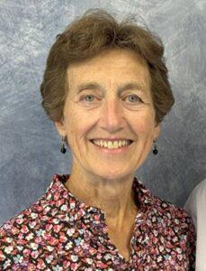 Portrait of a smiling woman with short brown hair wearing a collared shirt patterned with small red, pink, white, and blue flowers on a dark background. She has small drop earrings and stands before a textured gray‑blue backdrop, lit evenly for a professional headshot.