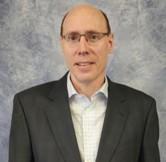Portrait of the Stewardship Deacon at Cheshire Lutheran Church, wearing glasses, a dark suit jacket, and a light checkered shirt, standing against a gray-blue studio backdrop.