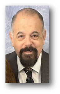 Portrait of the Fellowship Deacon at Cheshire Lutheran Church, wearing a dark suit jacket, white shirt, and patterned tie, smiling against a light blue backdrop.